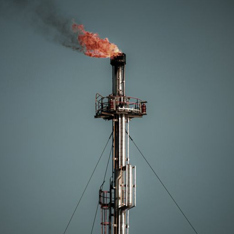 Industrial flare stack with a bright flame burning at the top against a clear sky, part of an oil/gas facility