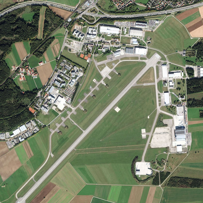 Aerial view of an airport with a long runway, terminal buildings, and surrounding green fields and roads.